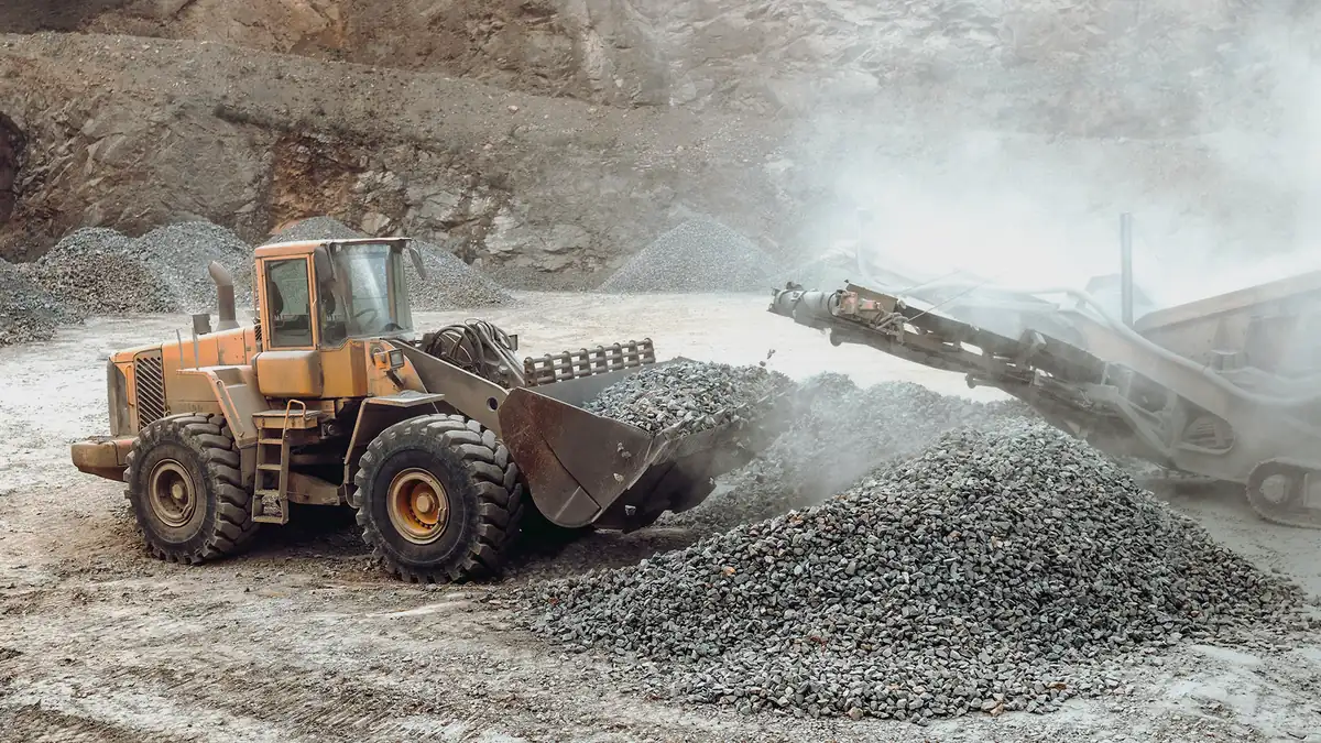 Wheel loader dumping crushed stone beside a conveyor at a dusty aggregates pit